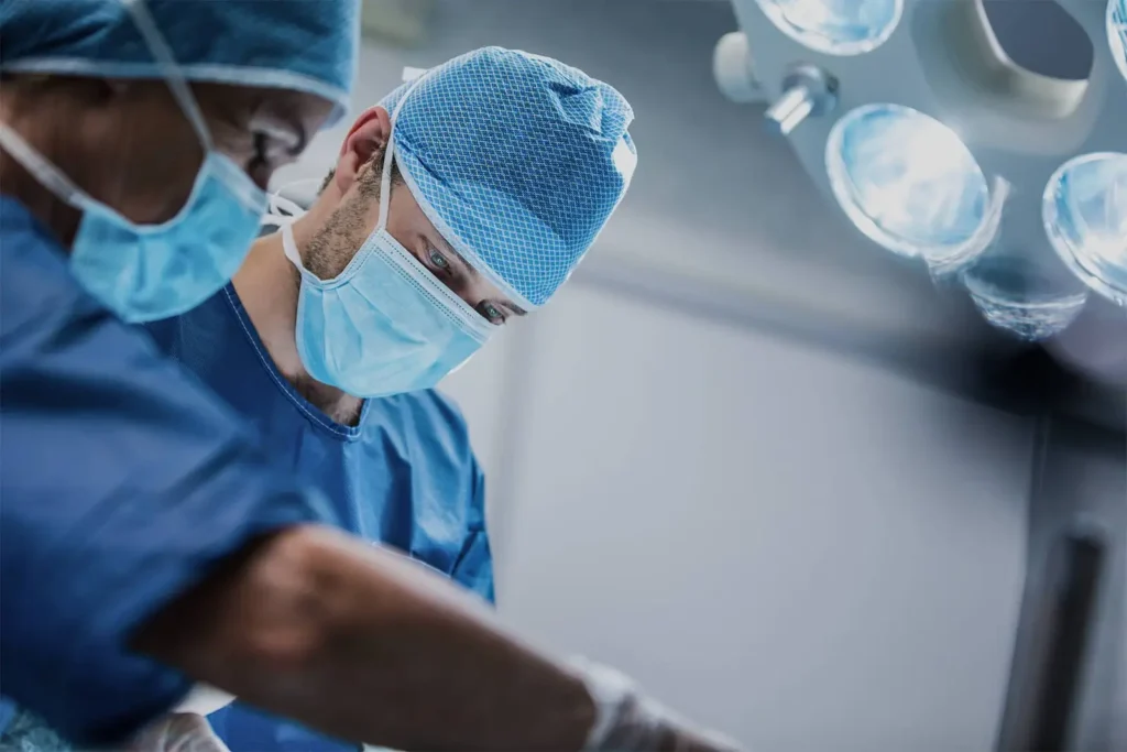 Dois cirurgiões paramentados com máscaras e toucas azuis realizando um procedimento cirúrgico com foco e precisão sob a luz de um foco cirúrgico no IOP.