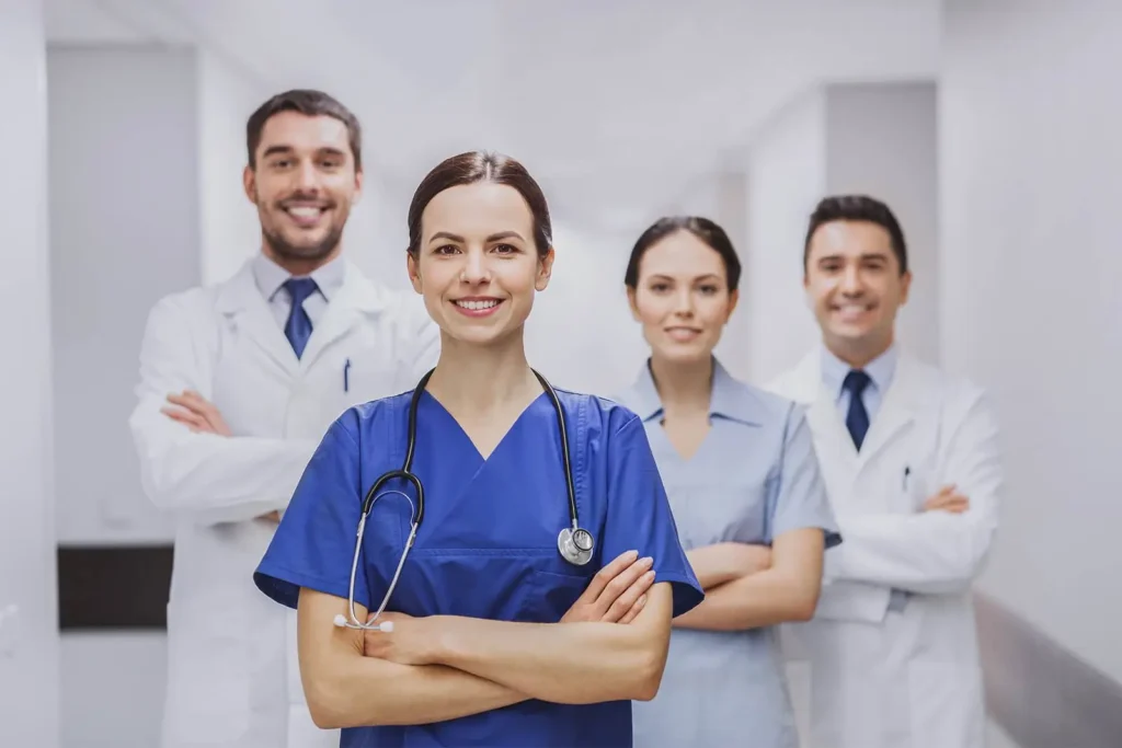 Médica sorrindo em destaque com braços cruzados e estetoscópio, liderando equipe de profissionais de saúde do IOP em um corredor hospitalar.