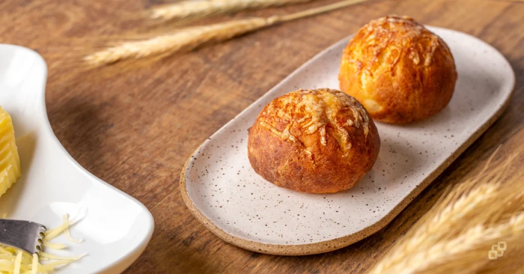 Pães de batata dourados e fofinhos servidos em um prato de cerâmica oval sobre uma mesa de madeira rústica, decorados com espigas de trigo seco ao fundo.