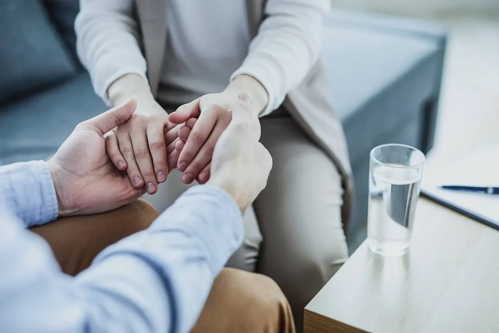 Plano detalhe das mãos de uma psicóloga segurando as mãos de um paciente em sinal de conforto e apoio durante uma consulta de terapia no IOP.