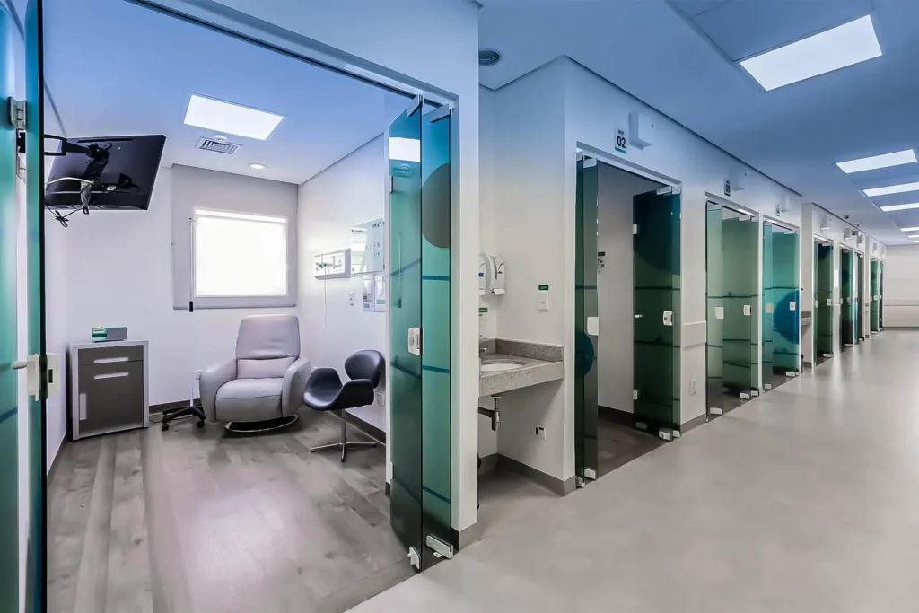 Corredor da ala de quimioterapia do IOP com cabines de tratamento individuais, poltronas reclináveis confortáveis e portas de vidro para privacidade do paciente.