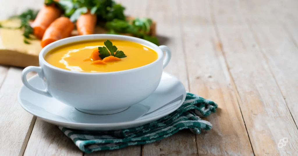 Tigela de porcelana branca com sopa creme de cenoura laranja, decorada com salsinha, sobre uma mesa de madeira rústica. Ao fundo, cenouras cruas com ramas, dentes de alho e um pote de creme compõem o cenário.